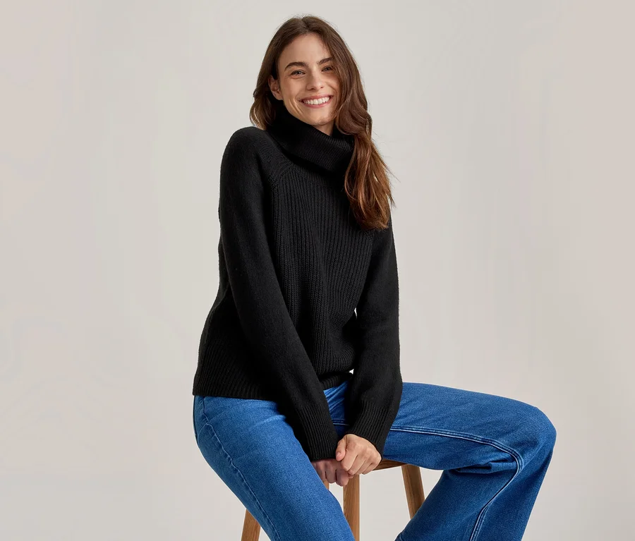 Lächelnde Frau mit langen braunen Haaren, die einen schwarzen Rollkragenpullover und blaue Jeans trägt und auf einem Holzhocker sitzt.