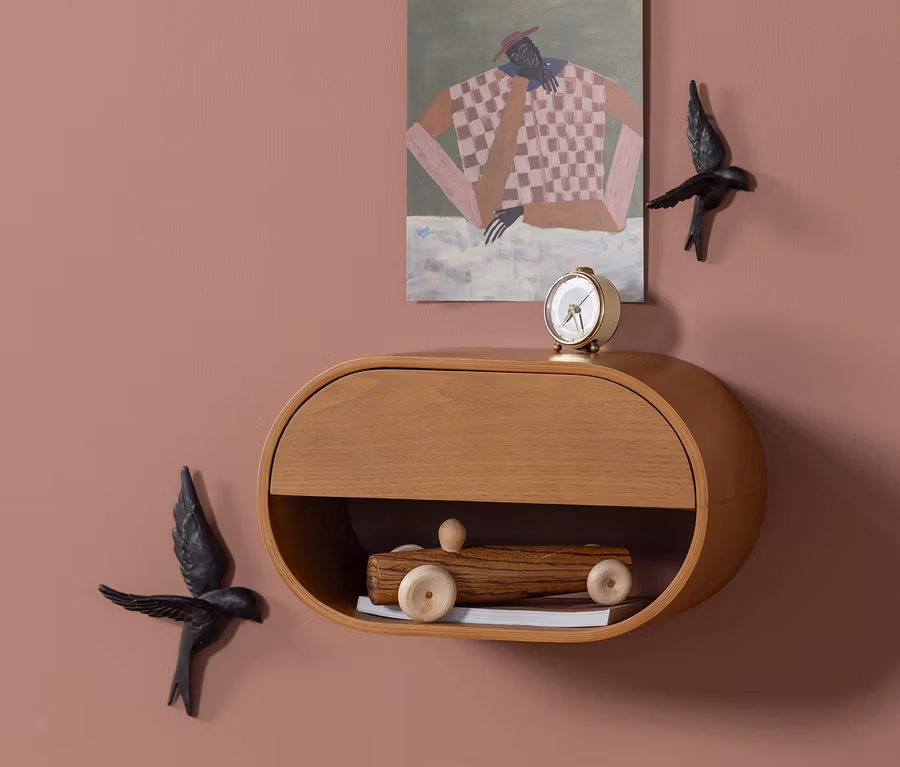Ein ovales Wandregal aus Holz mit einer Schublade und einem offenen Fach ist an einer altrosafarbenen Wand montiert. Eine kleine goldene Weckeruhr steht auf dem Regal, und ein hölzernes Spielzeugauto befindet sich im offenen Fach. Über dem Regal hängt ein Gemälde einer abstrakten Figur, flankiert von zwei schwarzen Vogel-Wanddekorationen.
