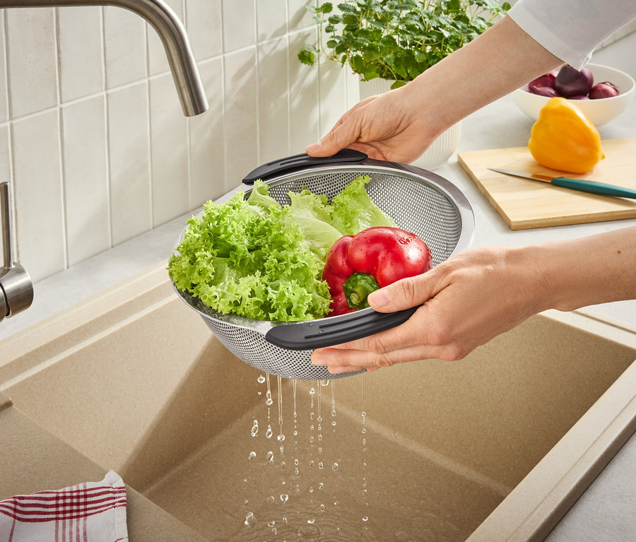 Hände spülen grünen Salat und eine rote Paprika in einem Metallsieb unter einem Wasserhahn, Wasser tropft in ein Spülbecken. Schneidebrett und weiteres Gemüse im Hintergrund.