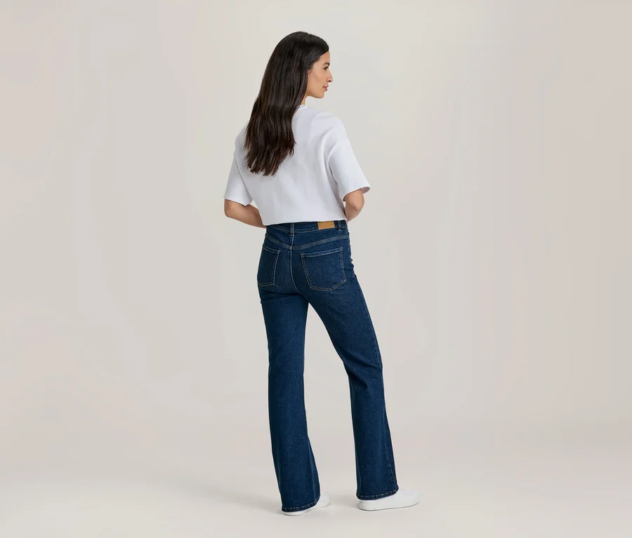 Frau von hinten, trägt ein weißes T-Shirt und eine dunkelblaue Schlagjeans, blickt nach rechts.