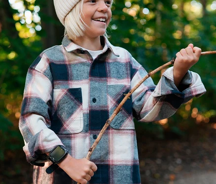 Lächelndes Kind in kariertem Hemd und Mütze hält einen Stock, trägt eine schwarze Xplora XGO2 Kids Watch am linken Handgelenk im Wald.