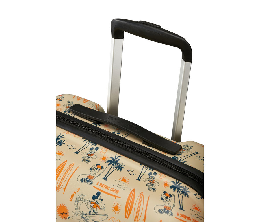 Nahaufnahme des oberen Teils eines beigen Koffers mit ausgezogenem silbernem Griff. Der Koffer zeigt ein Muster von surfendem Mickey Mouse, Palmen, orangen Sonnen und Surfbrettern, mit Text wie „SURFS UP“ und „A SURFING CHAMP“.