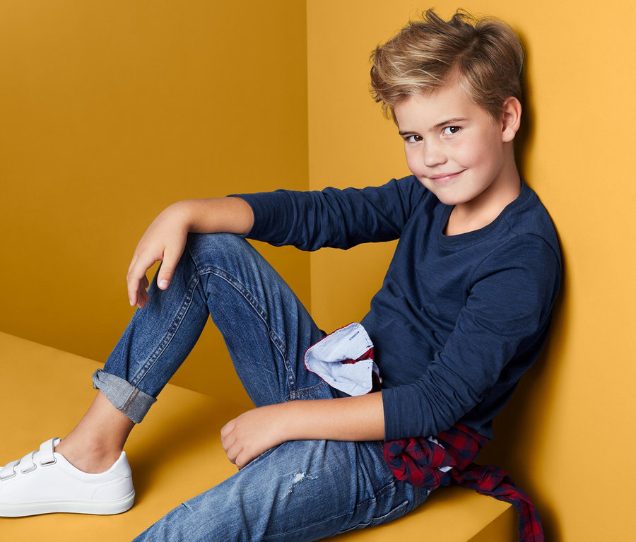 Ein Junge sitzt, trägt Jeans und ein blaues Langarmshirt (2er-Pack).