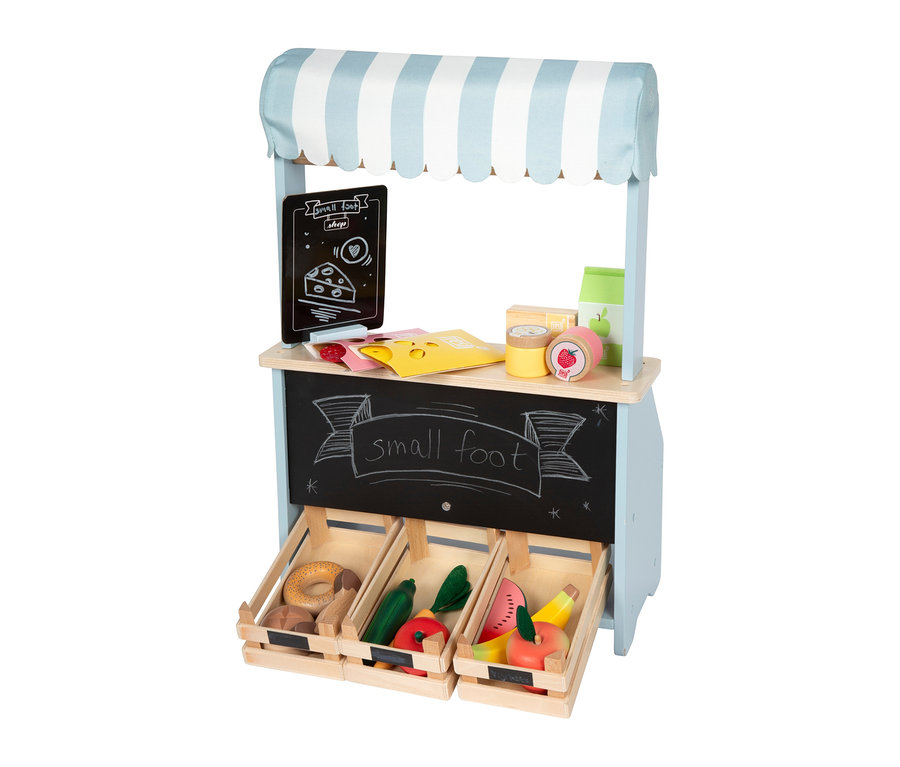 small foot kompakter Kaufladen mit blau-weiß gestreiftem Vordach. Die Holztheke zeigt Spielzeugkäse, Konserven und Obst, darunter drei Kisten mit Spielzeugbrot, Gemüse und Obst.