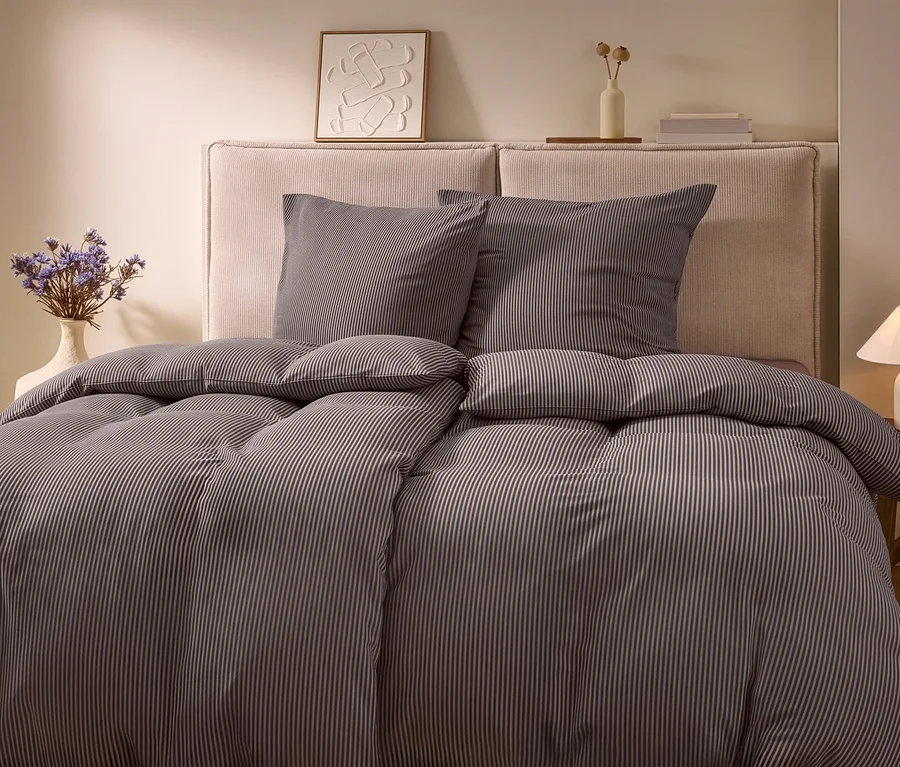 Ein Bett mit gestreifter Winterjersey-Bettwäsche, zwei Kissen und einem hellen Kopfteil. Links steht eine Vase mit Trockenblumen, rechts eine Lampe auf einem Holztisch.