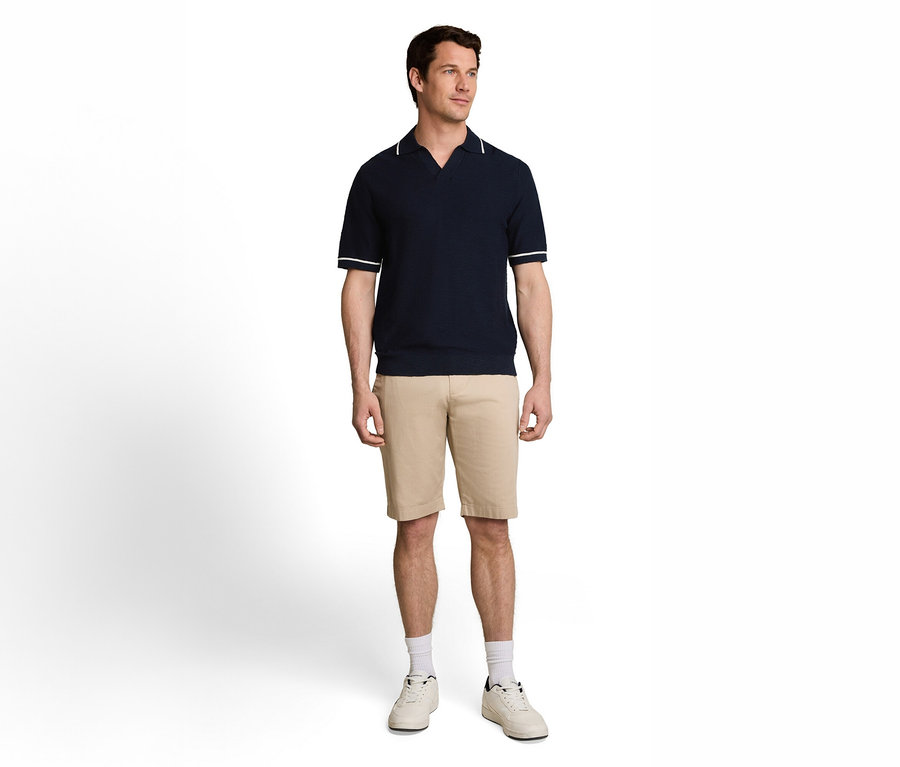 Ein Mann steht in einem dunkelblauen Poloshirt mit weißen Streifen, beigen Shorts und weißen Turnschuhen.