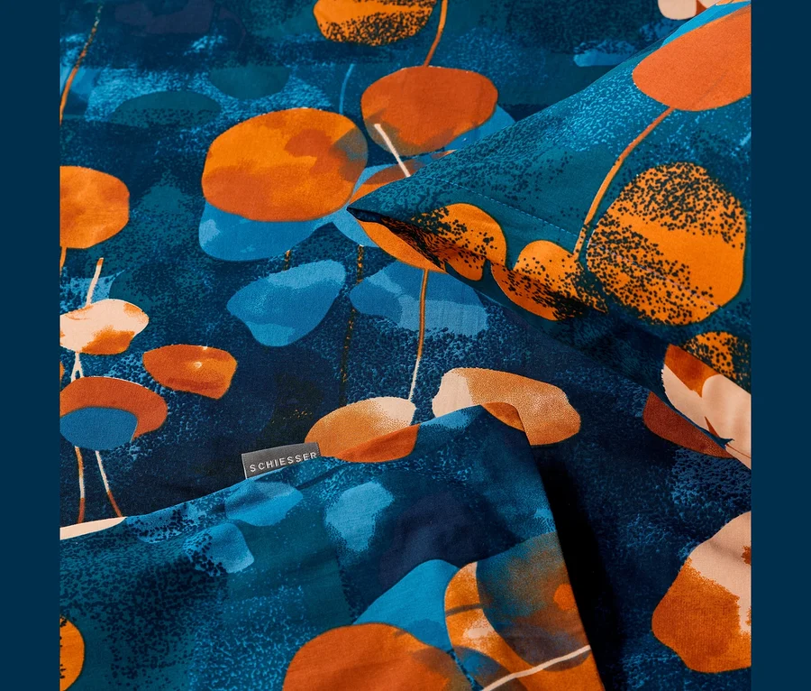 Nahaufnahme von SCHIESSER Renforcé-Bettwäsche mit einem abstrakten Muster aus orangefarbenen und blauen organischen Formen auf dunkelblauem Hintergrund. Ein schwarzes SCHIESSER Etikett ist sichtbar.