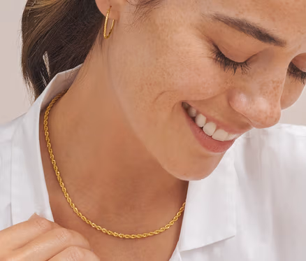 Eine lächelnde Frau schaut nach unten und berührt eine goldene Kordel Kette aus 925er Silber vergoldet. Sie trägt Gold-Creolen Basic flexibel und einen goldenen V-Ring.