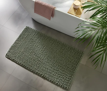 Mintfarbene benuta Badematte "Lynn" mit Noppenstruktur auf grauem Fliesenboden vor einer weißen Badewanne mit rosa Handtuch und einem Badewannenbrett.