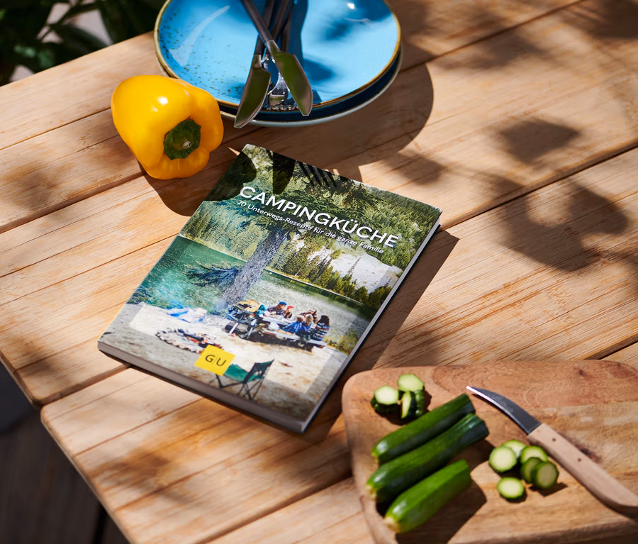 Auf einem Holztisch liegen das Buch "Campingküche", eine gelbe Paprika, blaue Teller mit Besteck und geschnittene Zucchini mit einem Messer.
