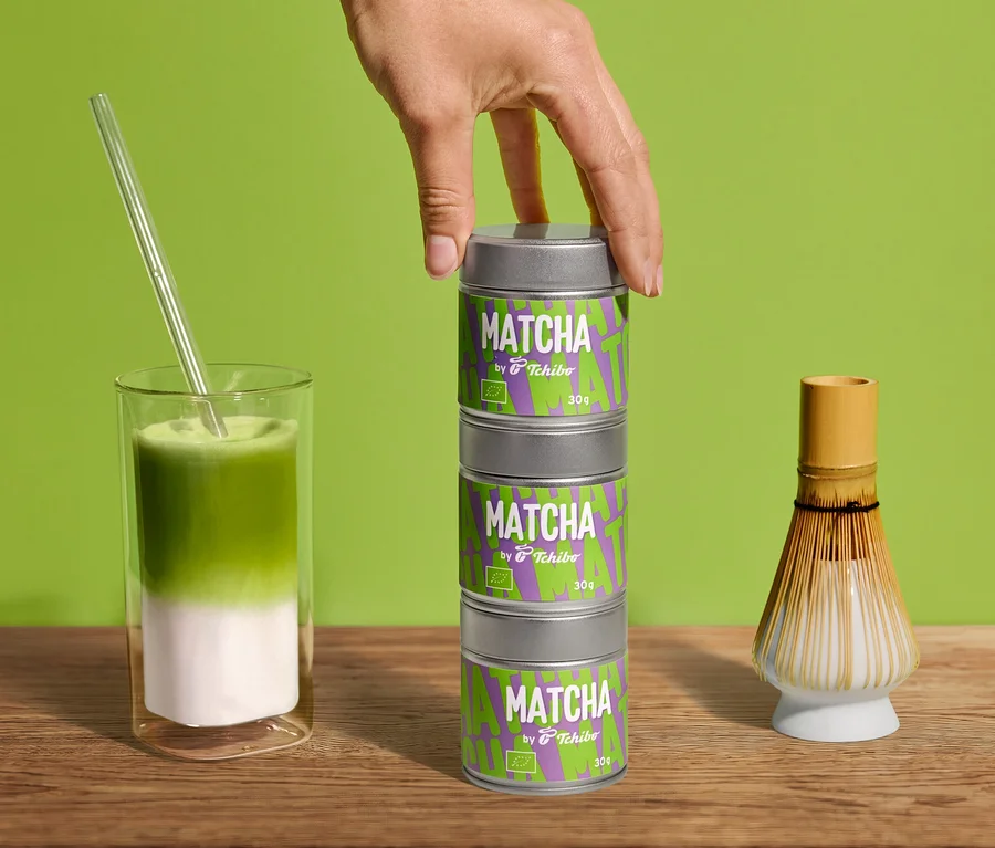 Hand stapelt drei Dosen Bio Matcha by Tchibo - 30 g. Matcha-Latte im Glas mit Strohhalm und Matcha-Besen auf Holz vor Grün.