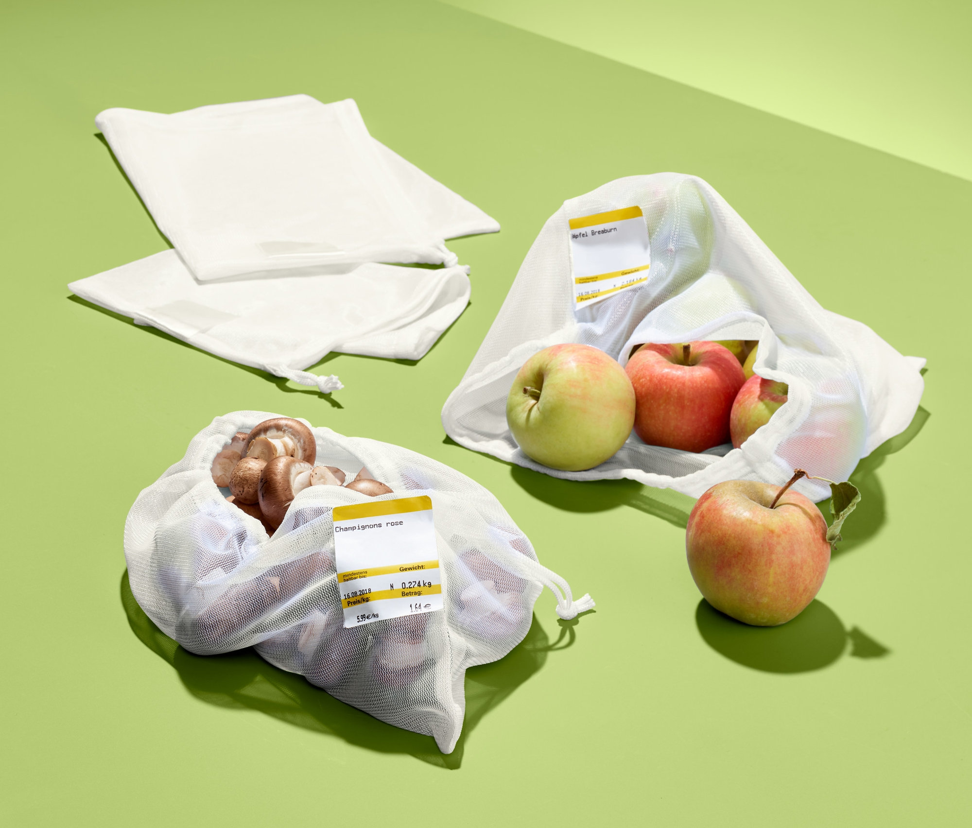 Grüner Hintergrund mit zwei weißen 4 Obst- und Gemüsenetzen, eines mit Champignons, eines mit Äpfeln.