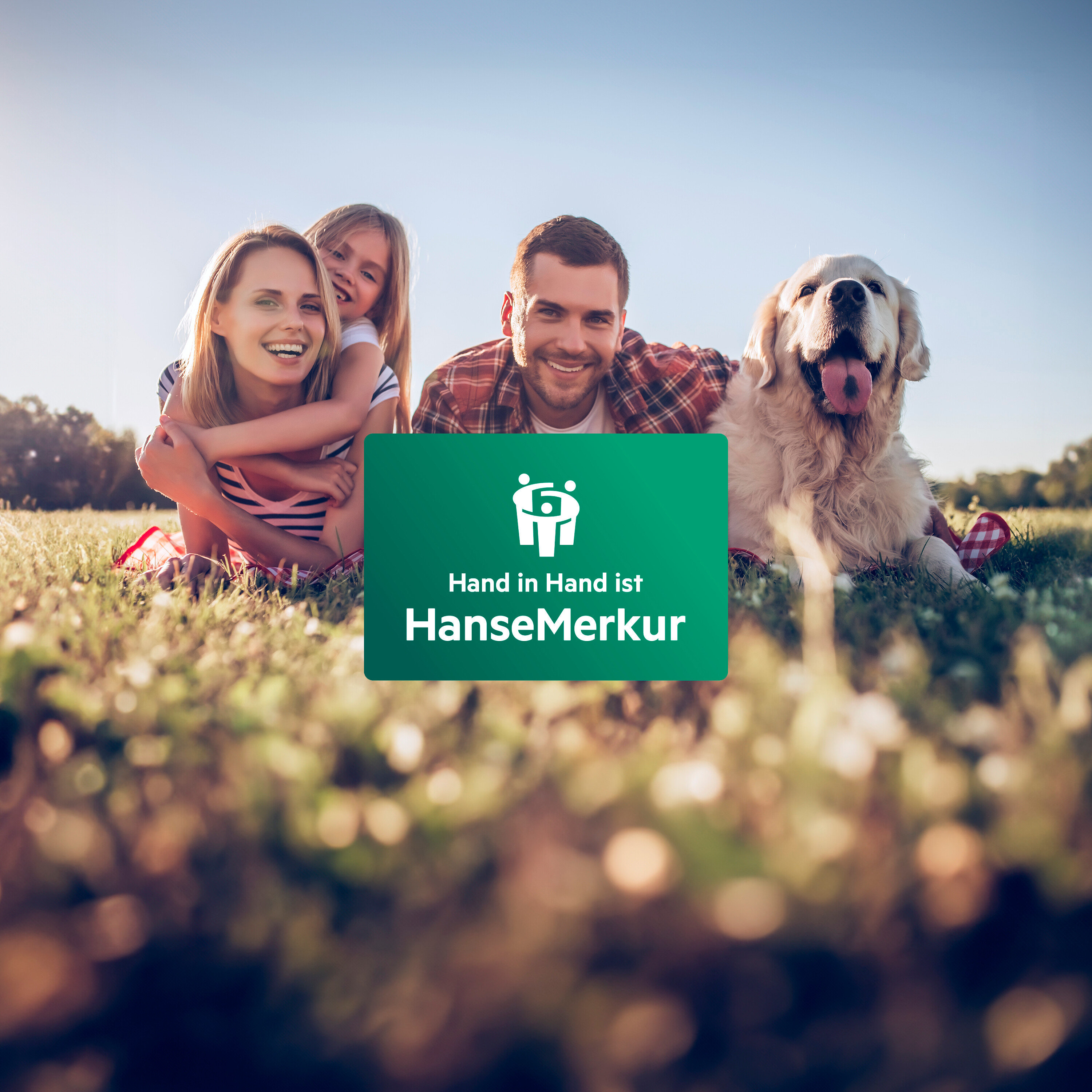 Eine lächelnde Familie mit Hund sitzt auf einer Wiese neben einem grünen Schild mit dem Logo und dem Text "Hand in Hand ist HanseMerkur".