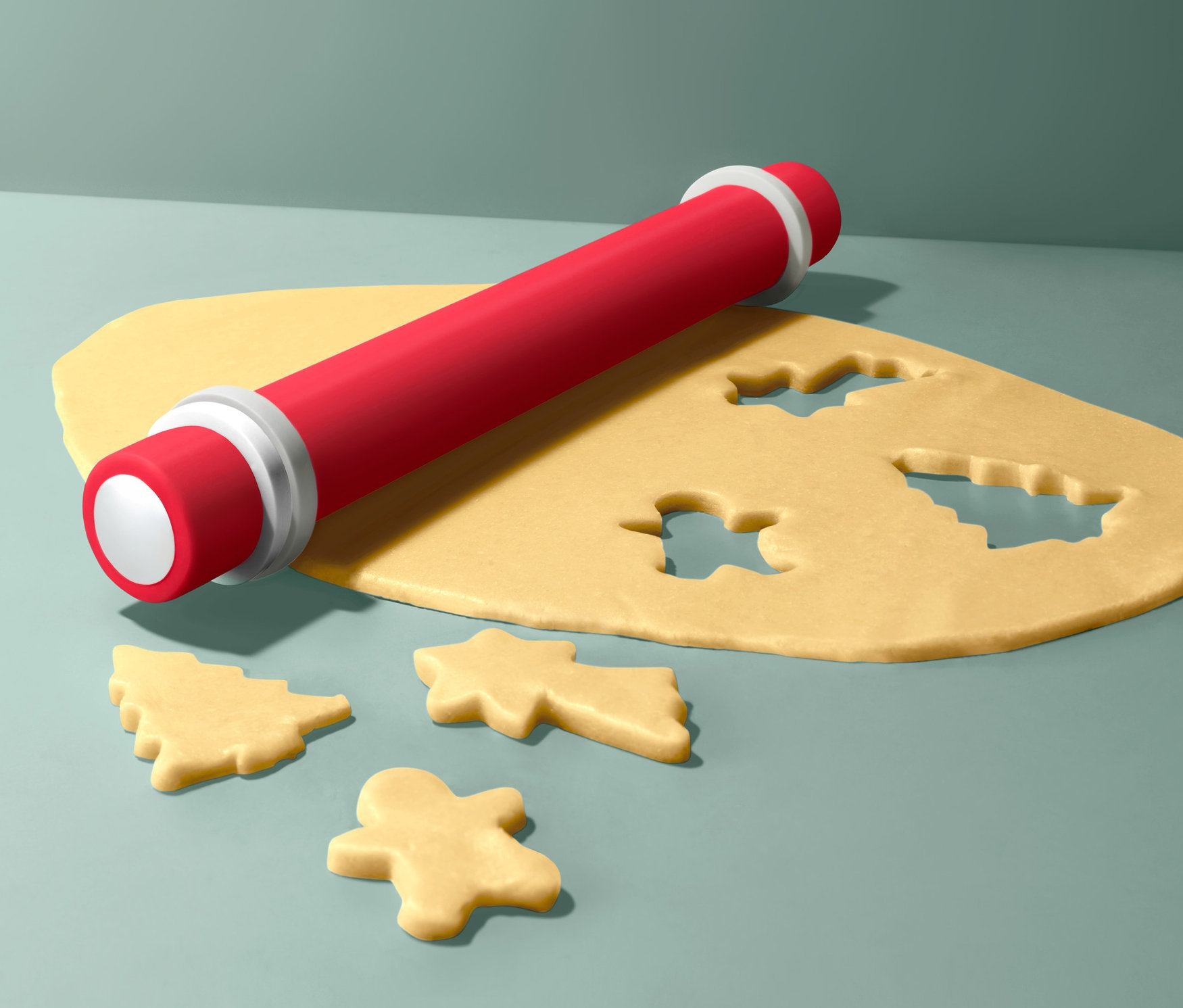 Eine rote Teigrolle liegt auf gelbem Teig mit ausgestochenen Formen von Weihnachtsbäumen und Lebkuchenmännchen.