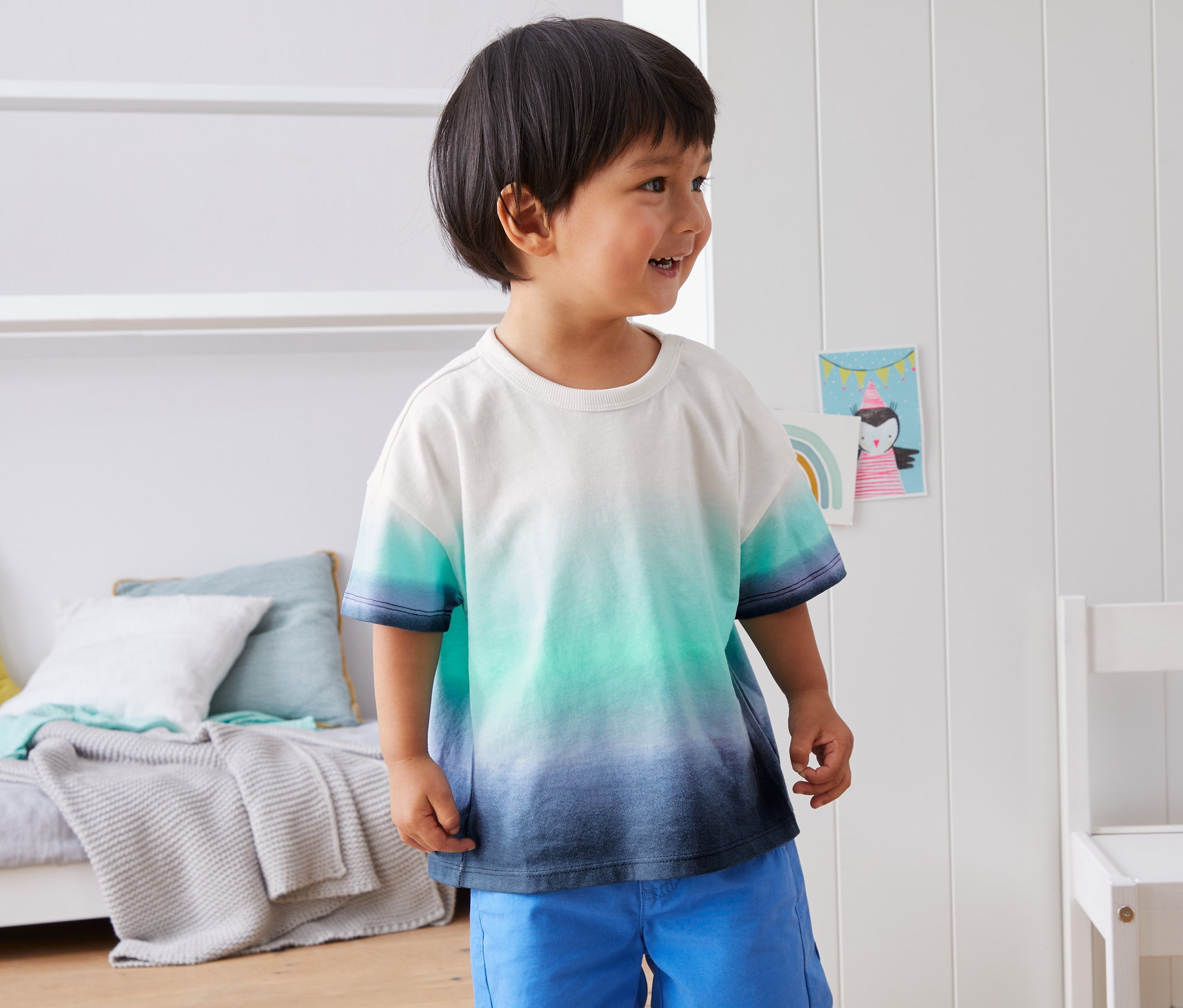 Kleiner Junge trägt ein T-Shirt aus Bio-Baumwolle mit Farbverlauf und blaue Shorts.