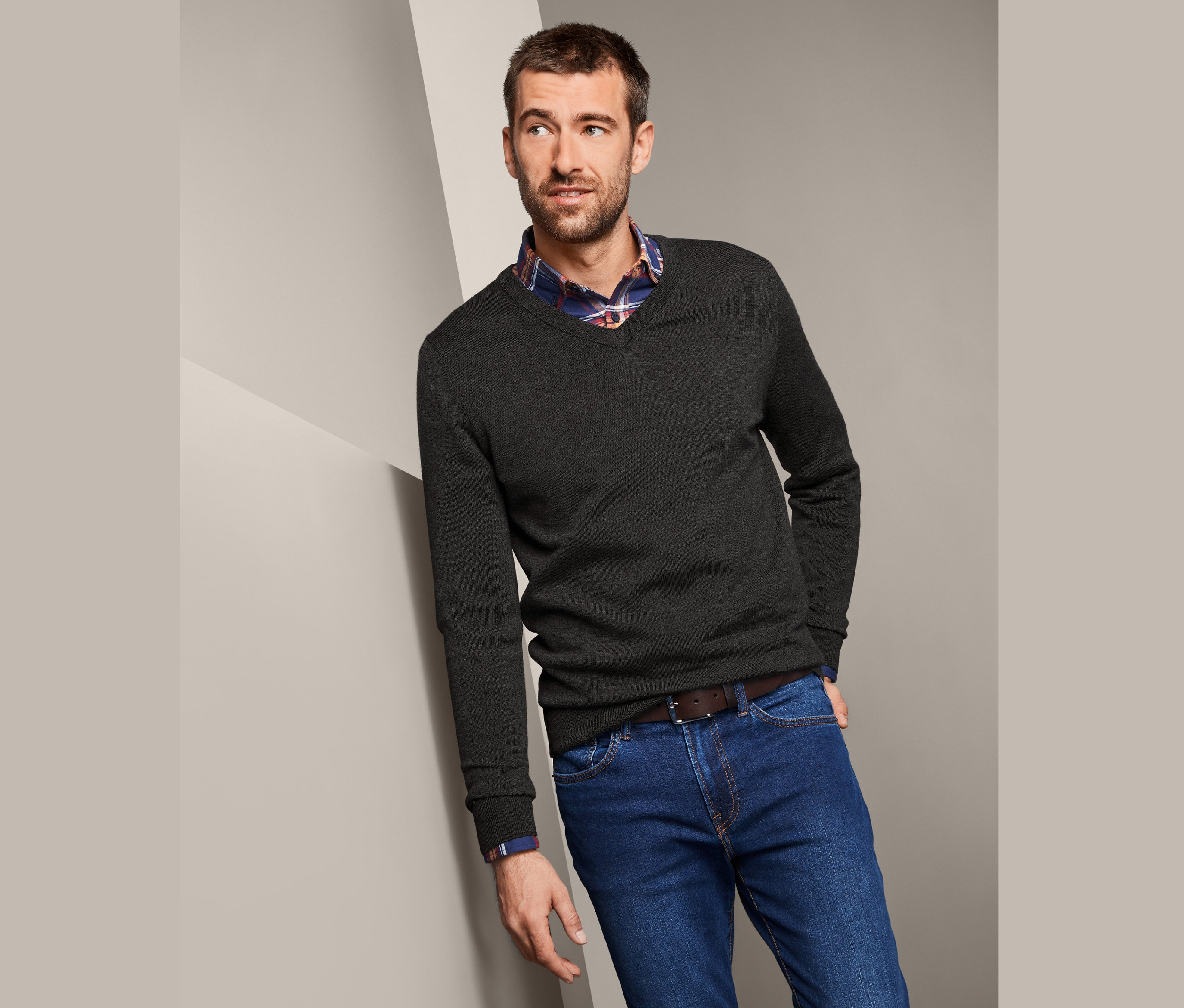 Mann lehnt an Wand, trägt Merino-Pullover, Hemd und Jeans.