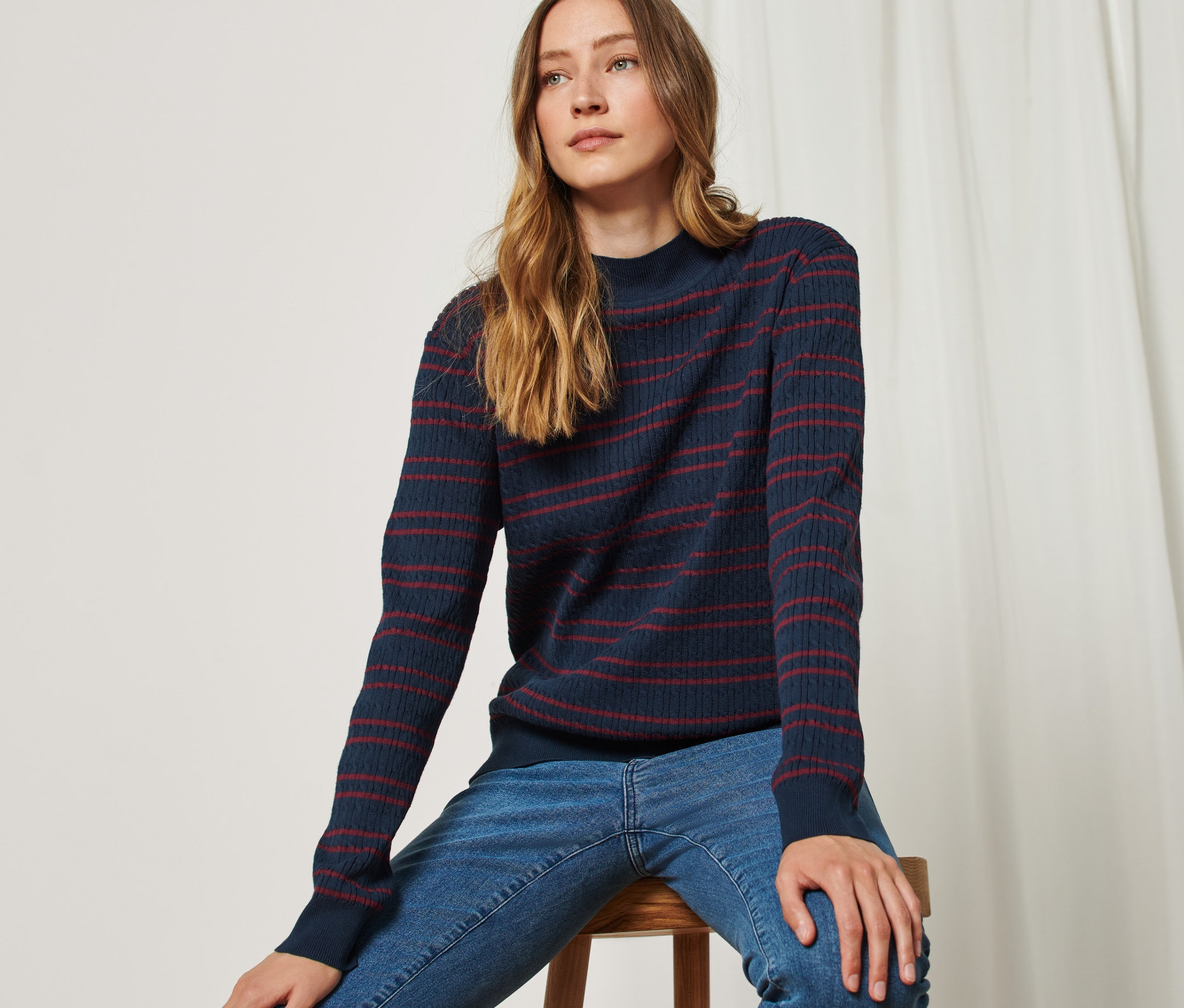 Frau sitzt auf einem Hocker und trägt einen blau-roten Feinstrickpullover mit Zopfmuster.