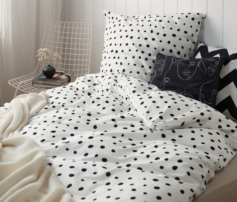 Ein Bett mit weißer Bettwäsche mit schwarzen Punkten, ergänzt durch dekorative Kissen und einer Strickdecke.