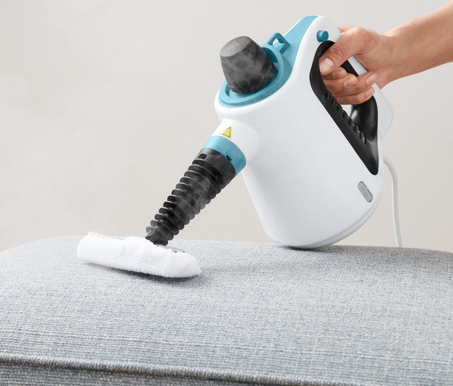 Eine Hand benutzt einen Dampfreiniger auf einem grau gepolsterten Hocker.