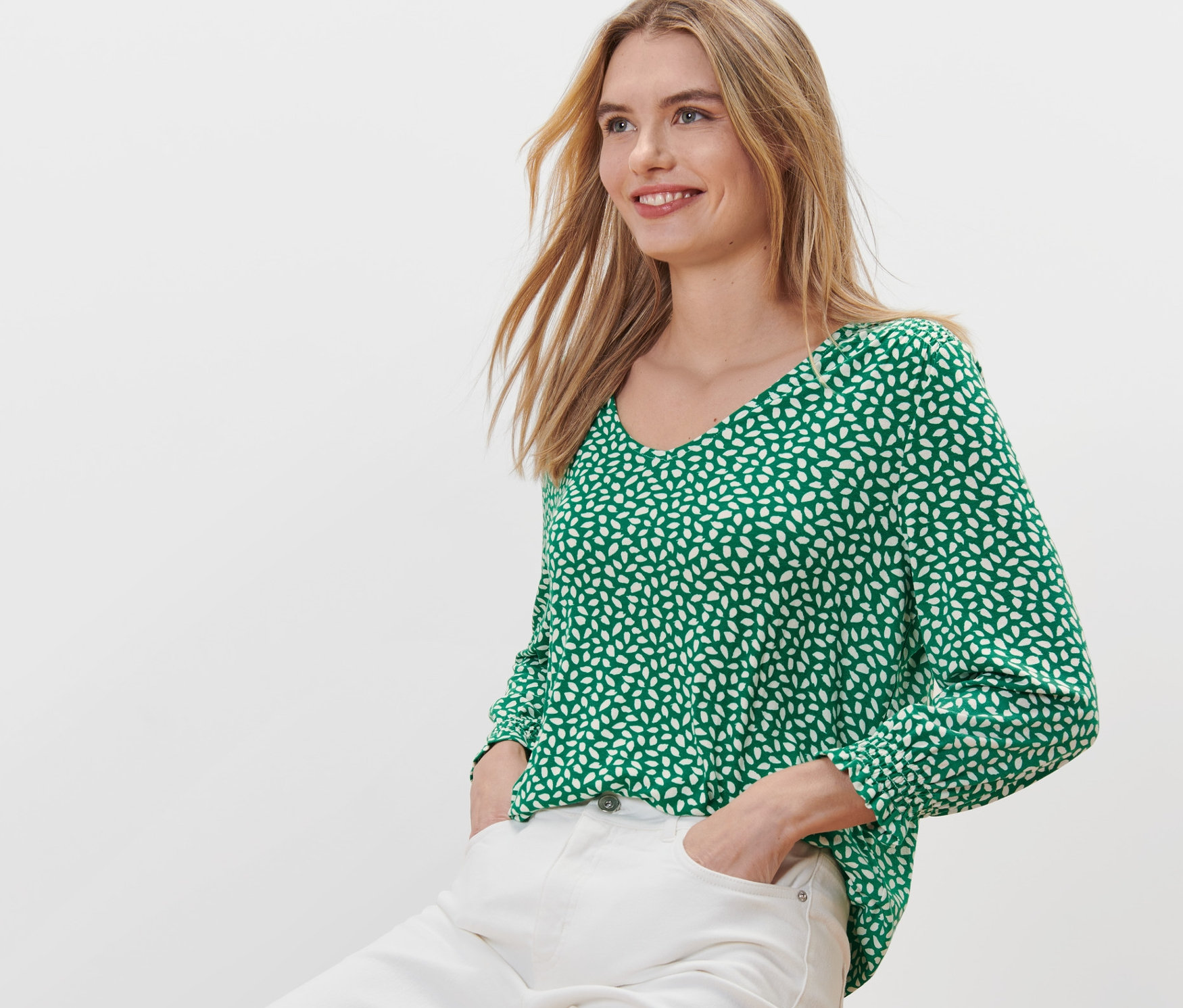 Frau sitzt auf einem Hocker und trägt ein grünes bedrucktes Blusenshirt.