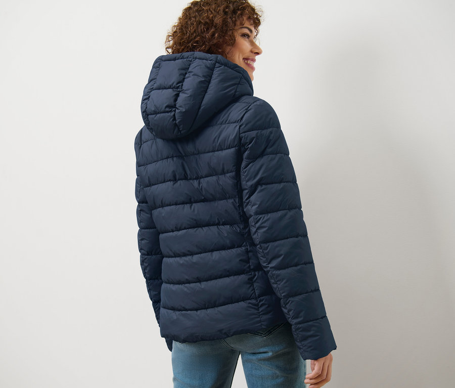 Frau mit lockigem Haar blickt über die Schulter und trägt eine dunkelblaue Steppjacke mit Kapuze.