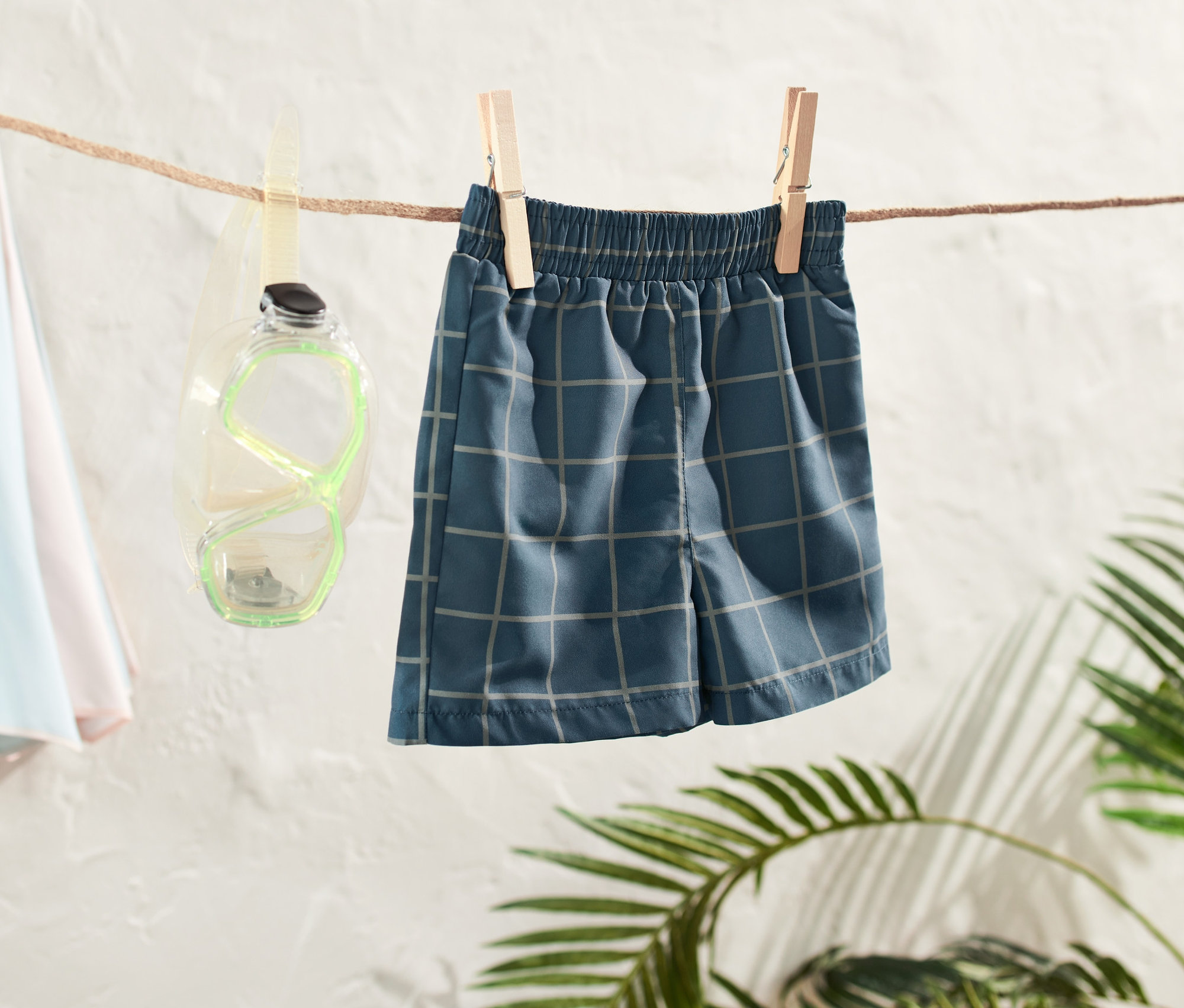 Kleinkind-Boardshorts hängen an einer Wäscheleine, daneben eine Taucherbrille und ein Strandschirm.