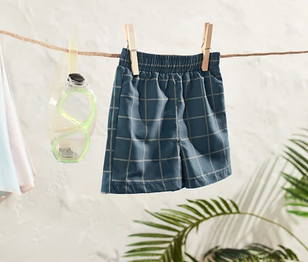 Kleinkind-Boardshorts hängen an einer Wäscheleine, daneben eine Taucherbrille und ein Strandschirm.
