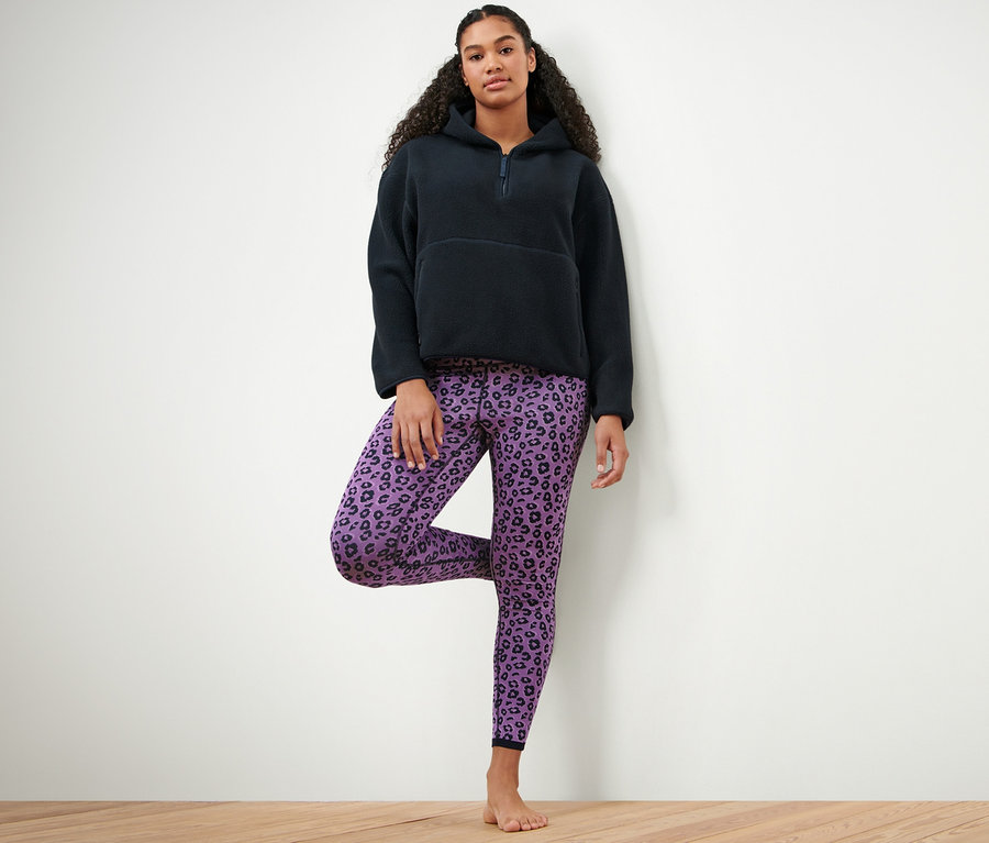 Frau steht auf einem Bein und trägt einen dunkelblauen Teddy-Kapuzenpullover und eine blaue und lavendelfarbene Wende-Sporttight.