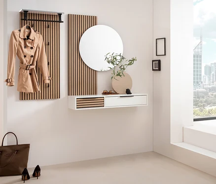 Eine moderne Garderobe mit einer weißen Konsole »Loft« und einem runden Spiegel »Loft« an der Wand. Ein beiger Trenchcoat hängt an einer Garderobenleiste. Rechts steht eine weiße Sitzbank unter einem großen Fenster mit Stadtblick.