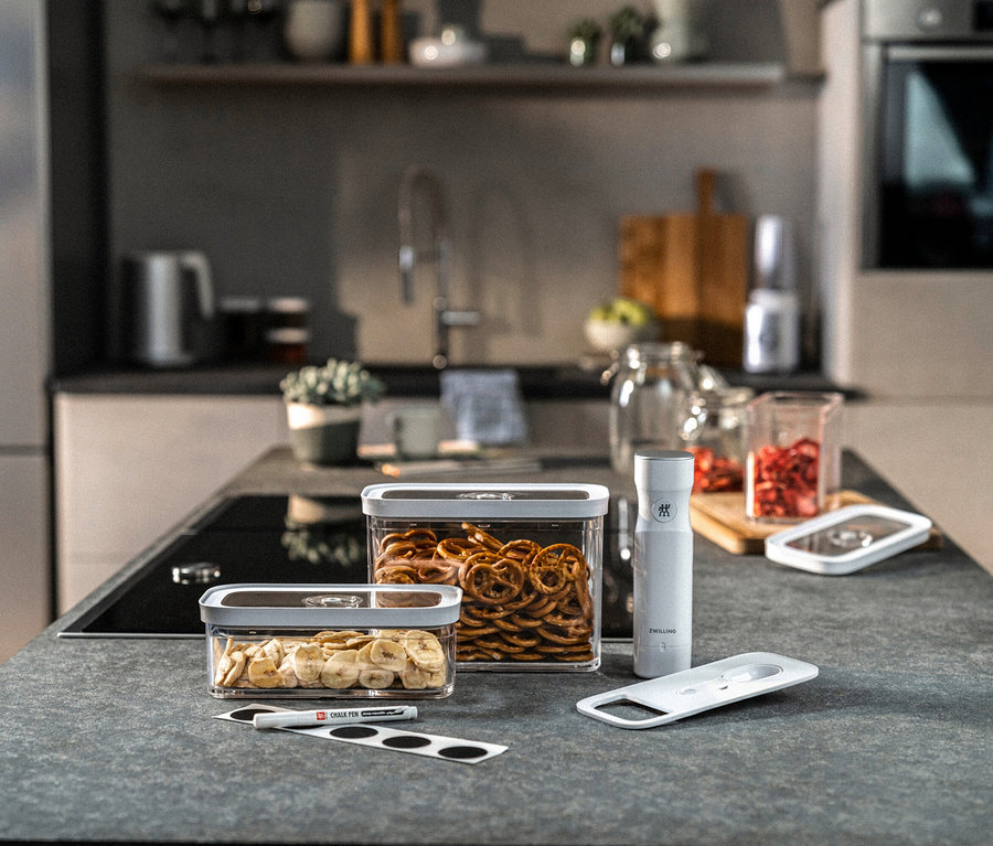 Zwei transparente Behälter mit Bananenchips und Brezeln, eine weiße Vakuumpumpe und ein Deckel des ZWILLING FRESH & SAVE CUBE Starter-Sets M auf einer grauen Küchentheke.