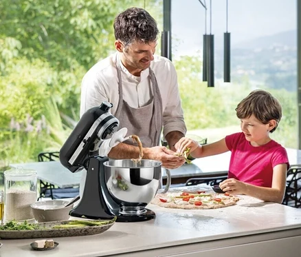 Ein Mann mit Schürze und ein Kind in einem pinkfarbenen T-Shirt bereiten Pizza zu und verwenden eine schwarze KitchenAid Küchenmaschine Artisan 5KSM125.