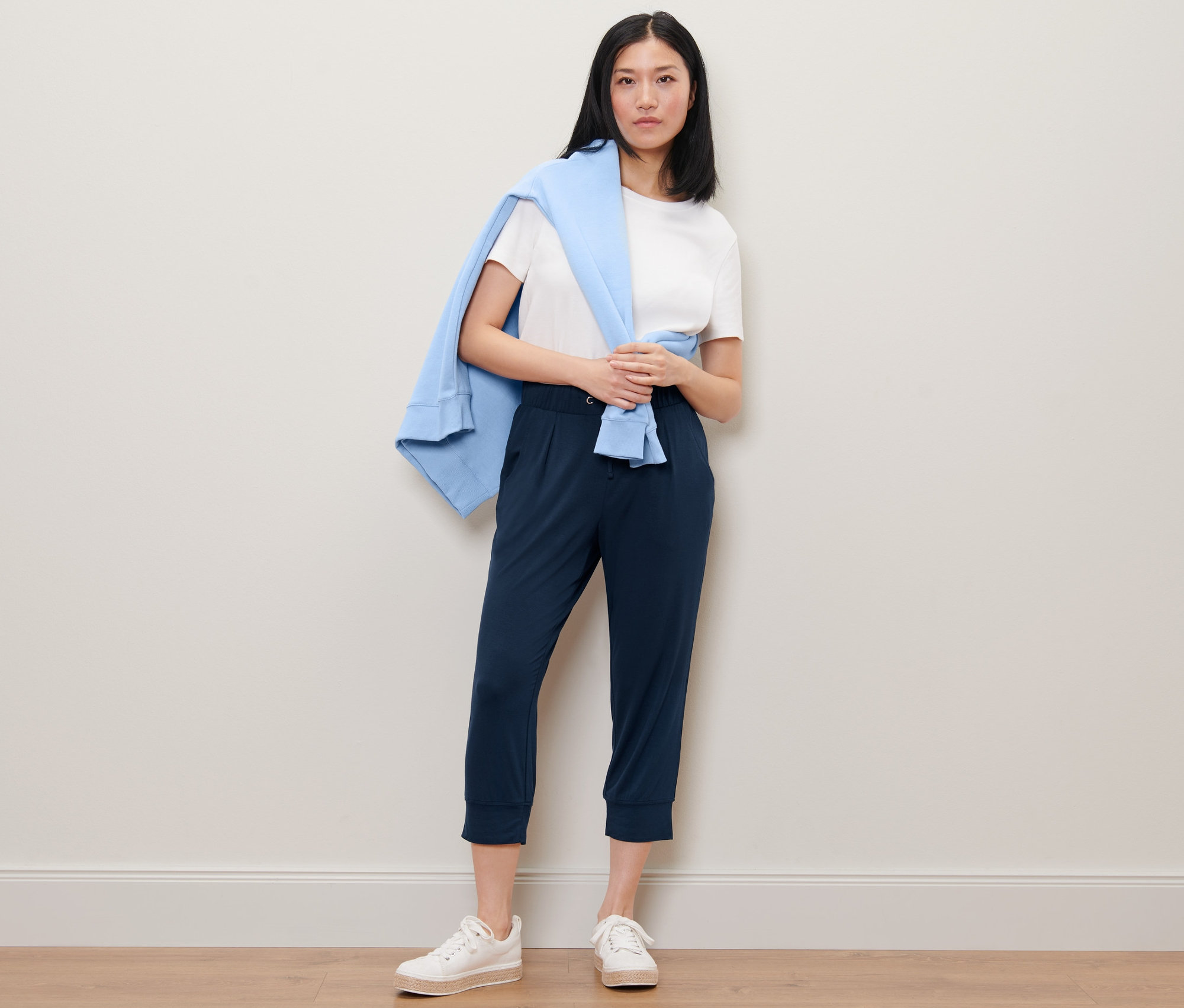 Eine Frau steht und trägt eine 3/4-Jerseyhose, ein weißes T-Shirt und einen blauen Pullover über den Schultern.