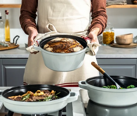 Mann in Schürze hält Brot in grauem Staub La Cocotte Kochtopf. Auf dem Herd stehen zwei weitere Kochtöpfe mit Speisen.