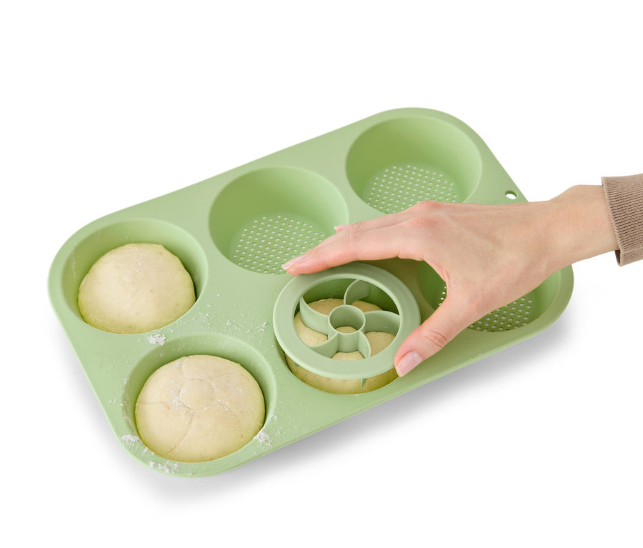 Eine Hand benutzt einen Ausstecher für Brötchen in einer hellgrünen perforierten Brötchenbackform, in der sich bereits zwei Brötchen befinden.