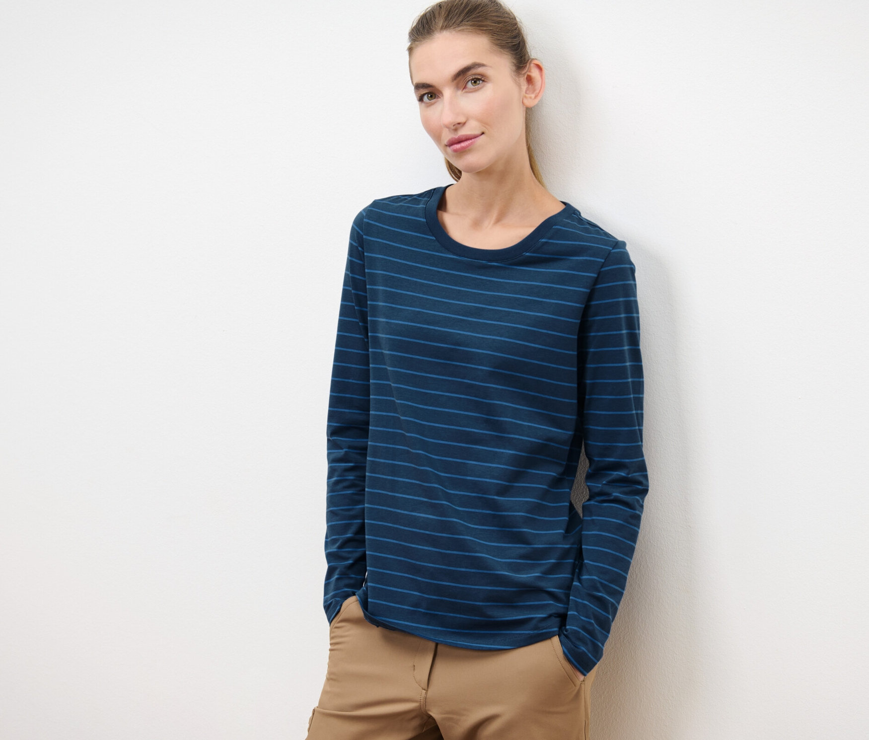 Frau lehnt an einer weißen Wand und trägt ein Langarm-Sportshirt und eine braune Hose.