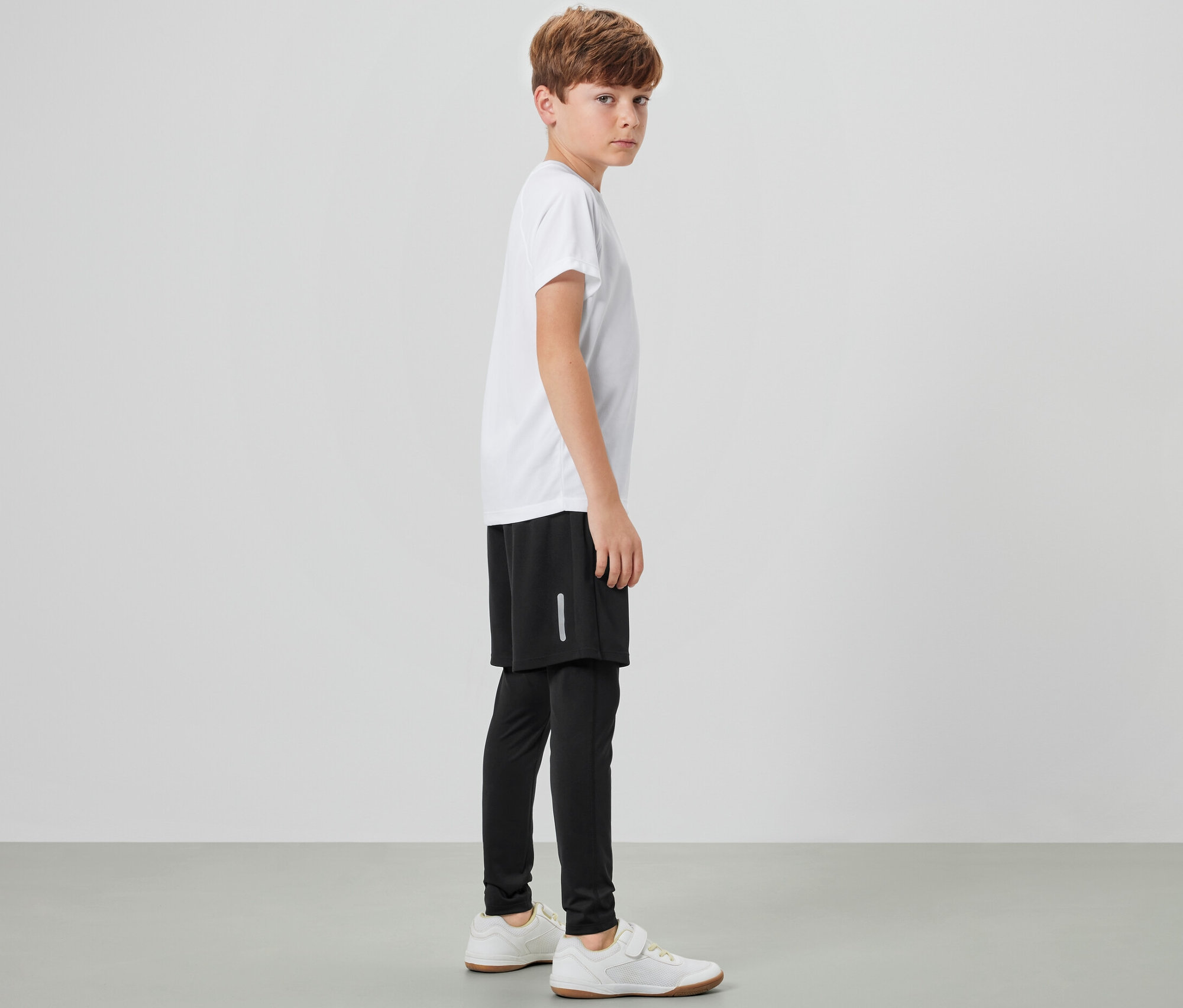 Ein Junge steht seitlich und trägt eine Kinder-Sporttight mit Shorts, ein weißes T-Shirt und Turnschuhe.