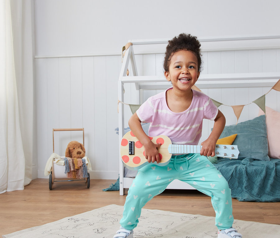 Lächelndes Mädchen spielt auf einer Spielzeug-Ukulele und trägt eine Schlupfhose aus Bio-Baumwolle.