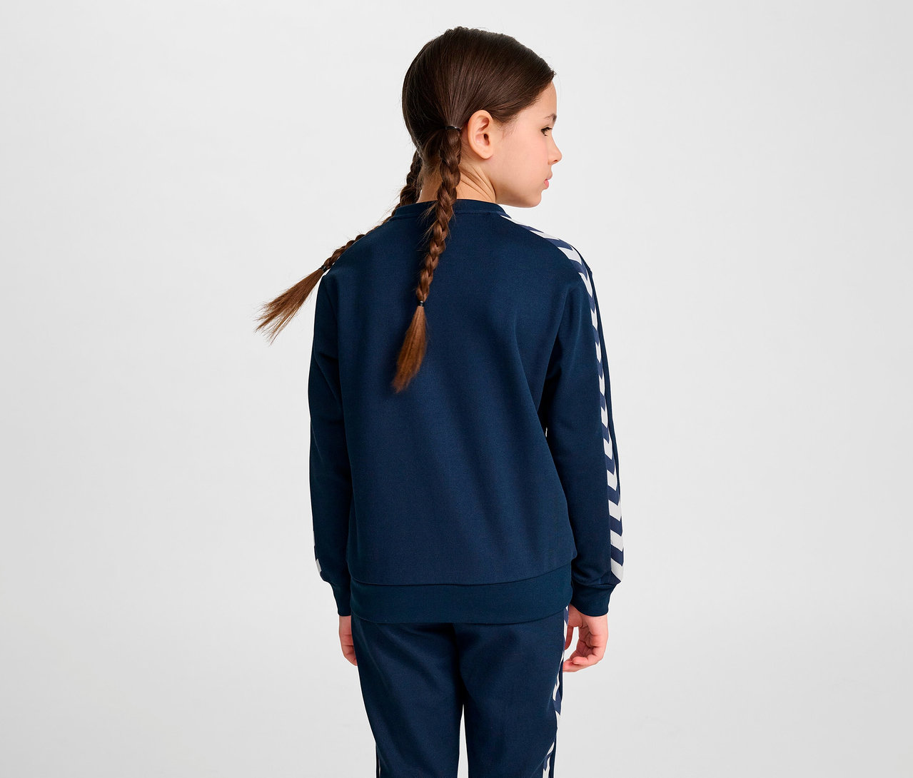 Mädchen mit Zöpfen steht mit dem Rücken zur Kamera und trägt eine blaue HUMMEL HMLJR Archive Reg Poly Zip Jacket.