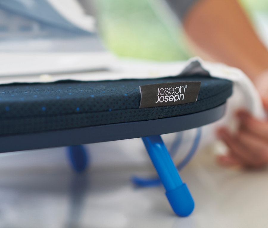 Detailaufnahme eines blauen Joseph Joseph Mini-Bügeltisches mit Markenetikett, mit einer Hand im Hintergrund.