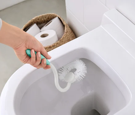 Frau putzt Toilette mit weißer Bürste mit türkisfarbenem Griff. Im Hintergrund befindet sich ein Korb mit Toilettenpapier.