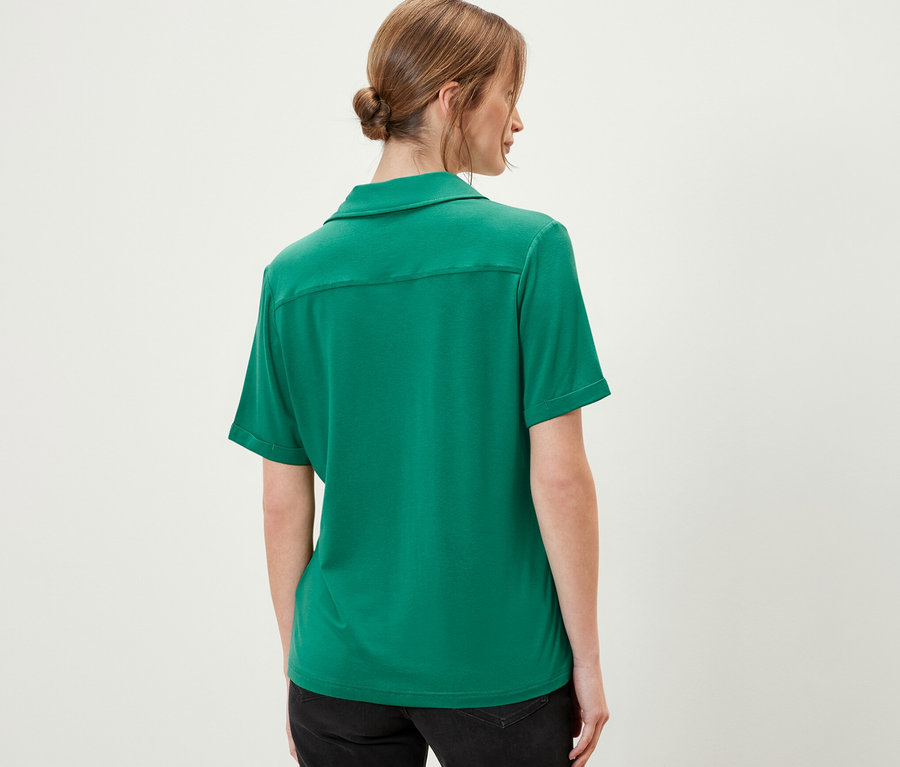 Rückansicht einer Frau, die ein smaragdgrünes Blusenshirt mit Knopfleiste und schwarze Shorts trägt.