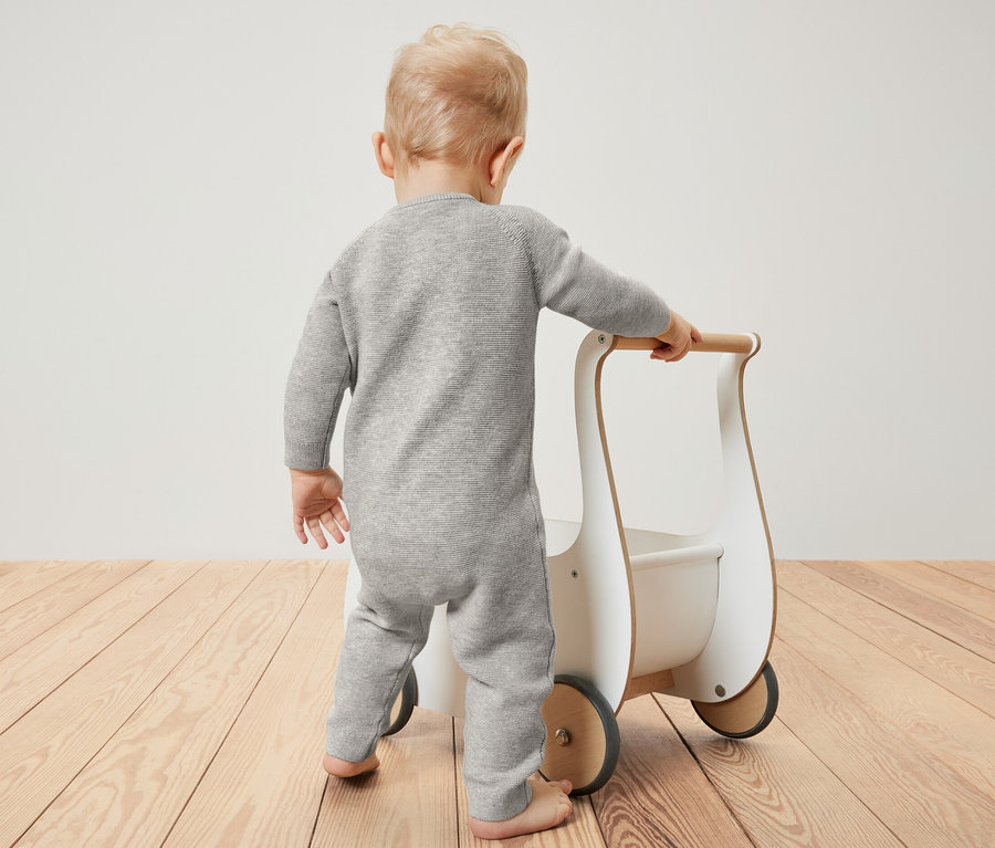 Baby im Strickoverall schiebt Lauflernwagen.