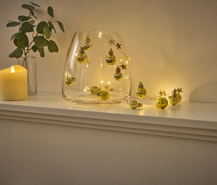 Auf einem weißen Regal stehen eine Glasvase mit LED-Lichtern in Sternform und goldenen Zierkugeln, daneben eine Glasvase mit Eukalyptus und eine gelbe Kerze.