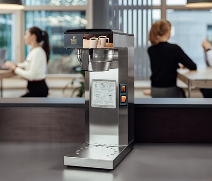Im Vordergrund befindet sich eine Tchibo Businessline Profi-Filterkaffeemaschine ohne Kanne, im Hintergrund sitzen Leute an Tischen.