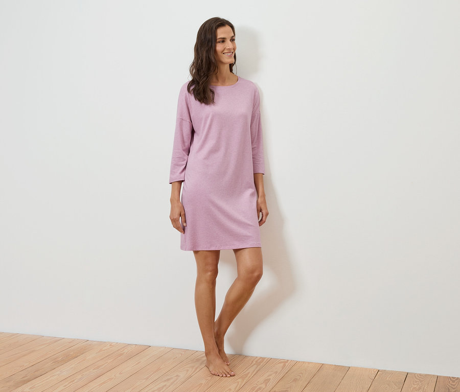 Eine Frau steht und lächelt, sie trägt ein lila Kleid mit Dreiviertelärmeln.