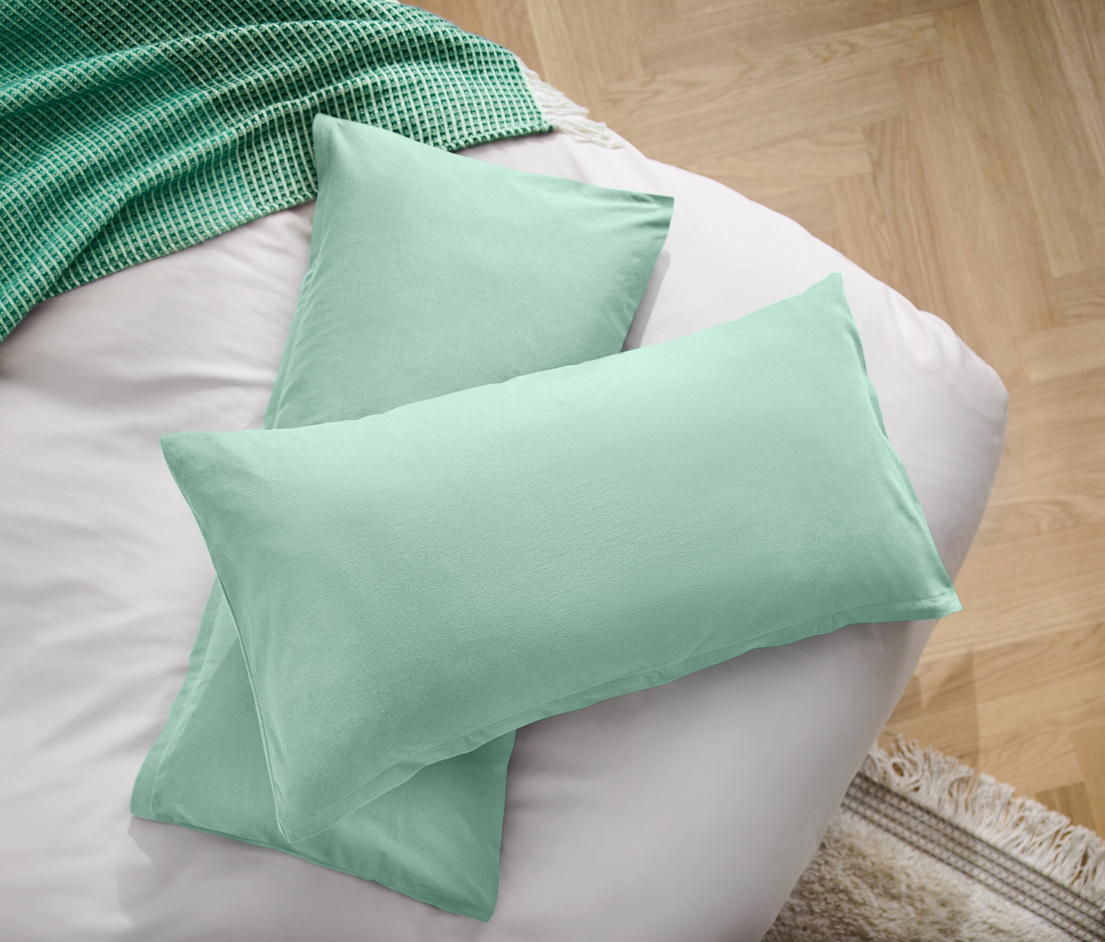 Zwei hellgrüne Kissen auf einem weißen Bettlaken neben einer grünen Decke.