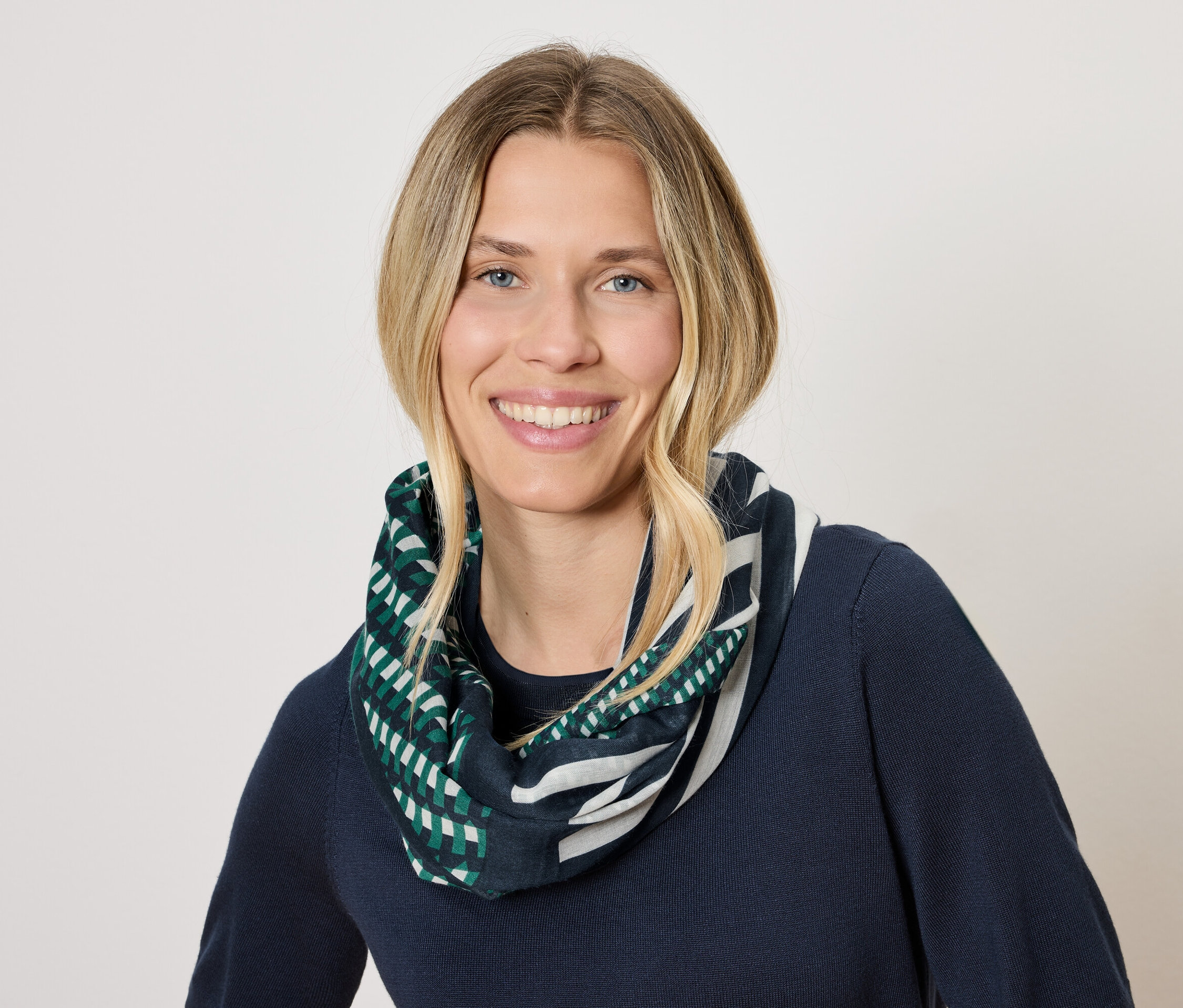 Frau mit blonden Haaren und einem Lächeln trägt einen dunkelblauen Pullover und einen Schal mit einem geometrischen Muster in Grün und Weiß.