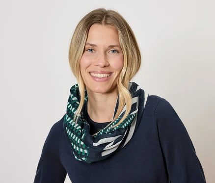 Frau mit blonden Haaren und einem Lächeln trägt einen dunkelblauen Pullover und einen Schal mit einem geometrischen Muster in Grün und Weiß.