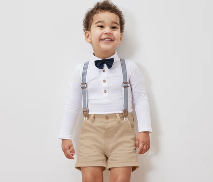 Lächelnder kleiner Junge in weißem Langarmhemd mit Fliege, Hosenträgern und Kinder-Shorts.