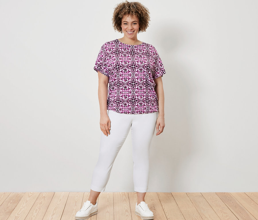 Frau posiert in einem bedruckten Blusenshirt in Berry-Farbe und einer weißen 7/8-Stretchhose.